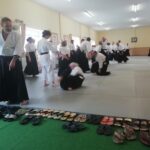 Lots of footwear lined up along the mat. One person pointing at them, looking confused