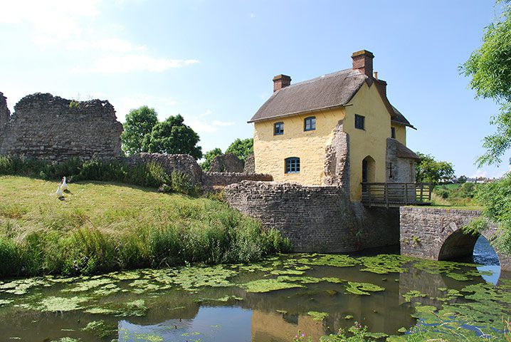 A yellow-washed, thatched, Somerset cottage by a footbridge over a small river.